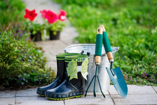 The image shows a garden setting with a paved pathway on which a pair of black rubber garden boots with a green and white leafy pattern are placed. Inside the boots, there are two small gardening tools with wooden handles and metal heads, one of which appears to be a hand trowel and the other a trans-planter. Next to the boots, there is a white plastic bucket, likely used for watering or carrying soil. Behind the bucket, a metal garden spade with a blue handle rests upright, partly leaning against the bucket. In the background, the garden features lush green grass, flowering plants with vibrant pink blooms, and further foliage, indicating a well-maintained outdoor space, typical of residential gardens in Mitcham area. The scene suggests preparations for gardening or planting activities, consistent with gardening services offered by Hedge Trimming Mitcham, in a natural outdoor environment under daylight conditions.