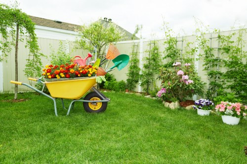 A well-maintained backyard garden in Mitcham featuring a lush, green lawn with evenly cut grass occupying the central area. To the right, a flower bed borders the garden, displaying blooming pink and purple flowers, with some potted plants in white containers placed nearby. Behind the flower bed, a white panel fence encloses the space, supporting climbing plants and small trees that add height and greenery to the garden's perimeter. In the foreground, a yellow wheelbarrow filled with colorful gardening tools and watering cans is positioned on the lawn, indicating recent or ongoing gardening activity. The background includes additional small trees and shrubs, contributing to the vibrant and welcoming outdoor environment. The area is bathed in natural light on a partly cloudy day, suggesting mild weather suitable for outdoor garden maintenance and landscaping services like hedge trimming, lawn care, and planting. This scene reflects a typical landscaped garden space in Mitcham, showcasing elements that a professional gardening service might care for or enhance. The image's clear, detailed composition and natural setting support local SEO signals for gardening and landscaping services in the Mitcham area.