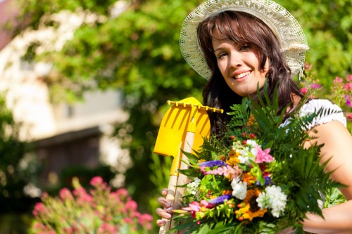 A woman in a white hat holding a large bouquet of colorful flowers and green foliage in a well-maintained garden. The garden features a neatly trimmed lawn with lush, dense grass in the foreground, bordered by flower beds containing various blooming plants with pink, purple, and white flowers. Behind her, a hedge forms a structured boundary, with tall trees and shrubs providing a natural backdrop. The environment appears sunny with clear natural light illuminating the scene, suggesting fair weather. The setting reflects a tidy, landscaped outdoor space suitable for gardening and outdoor maintenance, consistent with services offered by local gardening professionals such as Hedge Trimming Mitcham. The garden combines natural elements with careful design, showcasing mature greenery, vibrant blossoms, and clean paving or soil areas, creating a visually appealing outdoor environment.